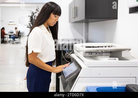 Side view of serious Asian young businesswoman in casuals with long hair using modern copier while working in creative office Stock Photo