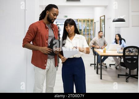 Multiracial young businessman holding digital tablet and listening to female colleague showing document while standing in creative office Stock Photo
