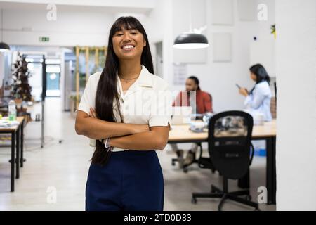 Portrait of smiling Asian young businesswoman in casuals with long hair standing with arms crossed in creative office Stock Photo