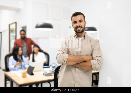 Portrait of smiling confident young Caucasian businessman in casuals with arms crossed leaning on wall in creative office Stock Photo