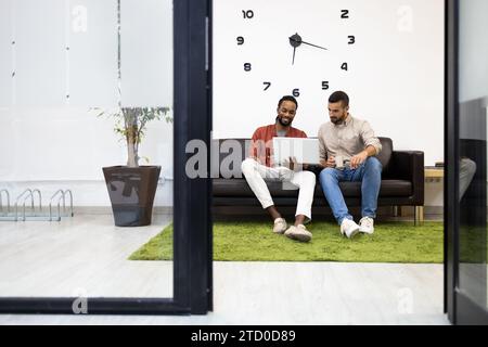 Multiracial male colleagues dressed in casuals discussing business plan over document and laptop while sitting on sofa against clock on wall in creati Stock Photo