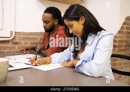 Young businesswoman sitting with male coworker and writing in document at desk while planning business strategy in creative office Stock Photo