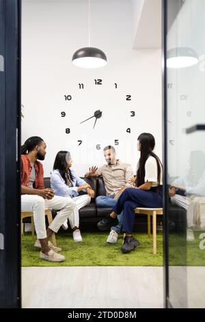 Full body of diverse colleagues discussing new project while sitting on sofa against clock on wall in lobby during meeting at creative office Stock Photo
