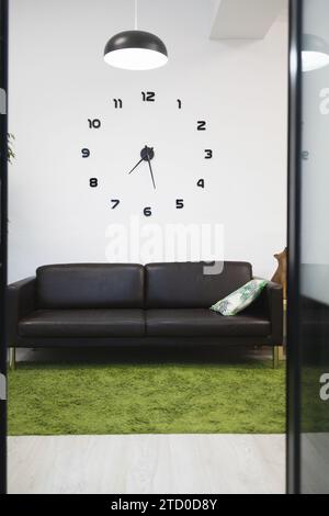 Creative black clock on white wall over empty sofa in lobby of coworking office seen through doorway Stock Photo