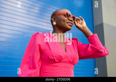 Low angle of confident African American female with shaved head in trendy pink outfit and sunglasses looking away while standing against blue wall Stock Photo