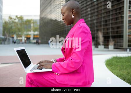 Side view of smiling African American female with shaved head entrepreneur in pink elegant suit sitting on street bench and typing on computer near bu Stock Photo