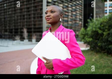 Smiling black businesswoman with shaved head wearing accessories in trendy pink suit holding laptop and looking away while standing on city street Stock Photo