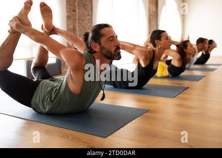 Group of adults in a yoga class performing the Dhanurasana bow pose, focusing on form and balance on their mats. Stock Photo