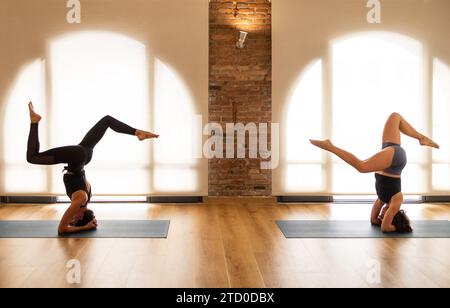 Two women practicing headstand yoga poses on mats in a sunlit room with arched windows and a brick wall. Stock Photo