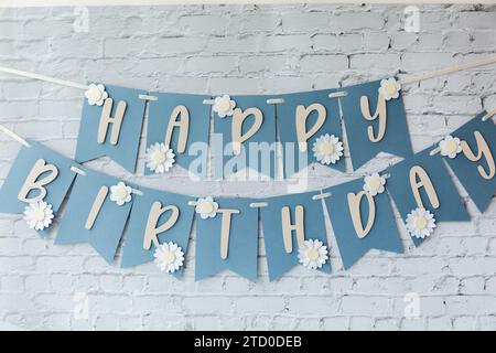 A cheerful blue banner spelling out "Happy Birthday" adorned with white daisy cutouts, set against a white brick background. Stock Photo