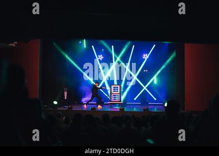 Magician in black suit performing magic trick with boxes on dark stage illuminated by blue and green spotlights in theater Stock Photo