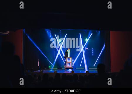 Magician in black suit performing magic trick with boxes on dark stage illuminated by blue and green spotlights in theater Stock Photo