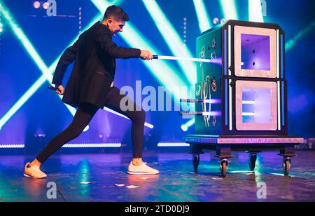 Side view of magician in black suit performing magic trick with boxes on dark stage illuminated by blue and green spotlights in theater Stock Photo