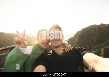 Portrait of smiling Caucasian young man taking selfie with cheerful woman showing victory sign while standing on top of mountain against clear sky Stock Photo