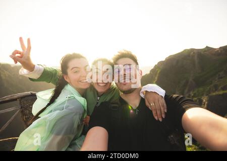 Portrait of cheerful Caucasian active young friends taking selfie looking at camera on mountain peak during sunrise at weekend hiking Stock Photo