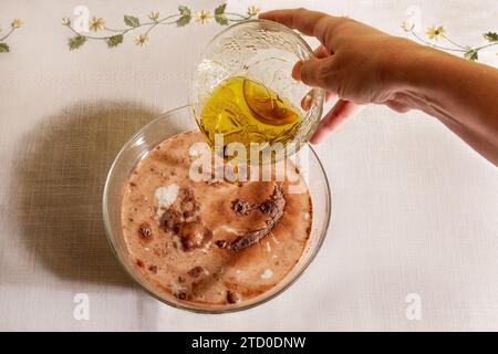 Top view of crop anonymous person is shown drizzling olive oil onto clear glass bowl filled with milk and powdered chocolate, on a table with embroide Stock Photo