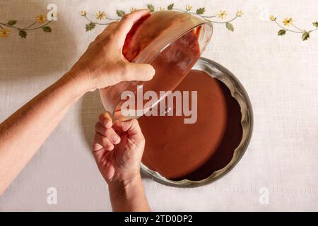 Top view of Hands of anonymous person carefully pouring smooth chocolate batter from a glass bowl into a round baking tray on a table Stock Photo