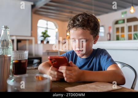 Cute focused boy text messaging over smart phone and sitting at dining table with drink in glass while spending leisure time in cafe Stock Photo