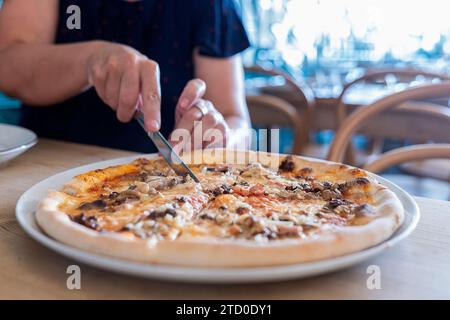 Crop anonymous person cutting pizza into slices served in plate with knife on wooden table while spending leisure time in restaurant Stock Photo