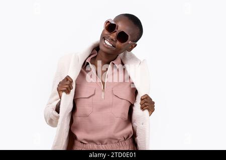 Cheerful young African American female in trendy long coat and sunglasses looking at camera while standing on white background Stock Photo