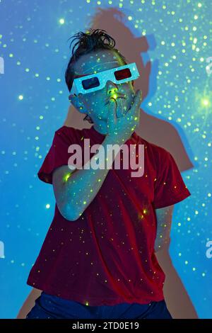 Confident boy in casual clothes with beads on face and 3D glasses with hand on chin standing against wall with illuminated neon lights in studio while Stock Photo