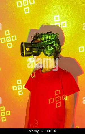 Boy in casual clothes and modern VR goggles experiencing virtual reality in illuminated studio with patterned neon lights Stock Photo