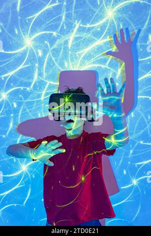 Amazed boy wearing virtual reality goggles with raised arms and open mouth experiencing cyberspace while standing in studio with neon lights Stock Photo