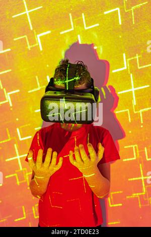 Boy in casual clothes and modern VR goggles experiencing virtual reality in illuminated studio with patterned neon lights Stock Photo