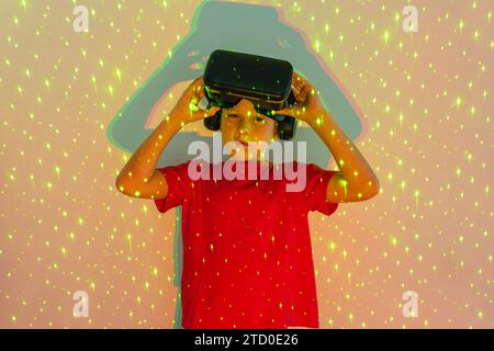 Portrait of Cute boy in casual clothes wearing virtual reality headset looking at camera while standing in patterned neon lights in studio Stock Photo