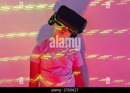 Girl in casual clothes and virtual reality headset experiencing cyberspace digital world in illuminated studio with neon lights Stock Photo