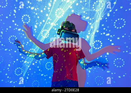 Boy in casual clothes and modern VR goggles experiencing virtual reality in illuminated studio with patterned neon lights Stock Photo