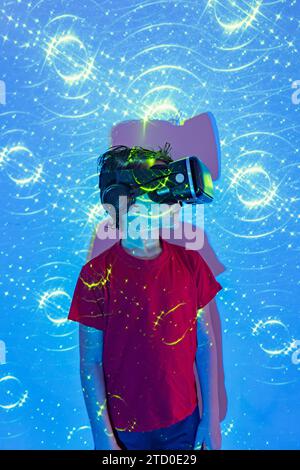 Boy in casual clothes and modern VR goggles experiencing virtual reality in illuminated studio with patterned neon lights Stock Photo