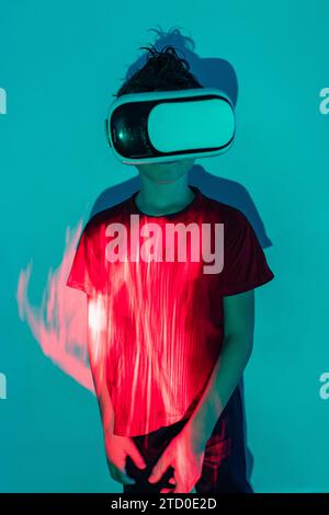 Boy in casual clothes and modern VR goggles experiencing virtual reality against blue wall with neon lights Stock Photo