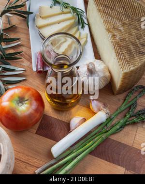 A variety of fresh ingredients including artisan cheese, olive oil, tomatoes, and asparagus laid out on a wooden cutting board. Stock Photo
