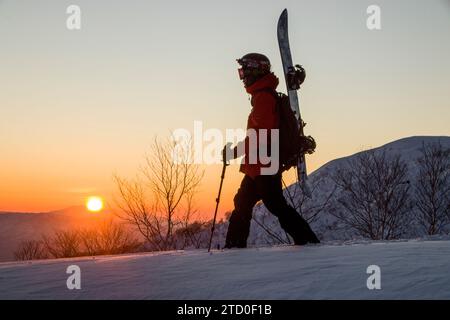 Side view of active anonymous man in sportswear with skies and poles walking on snow covered mountain against clear sky and beautiful sun during sunse Stock Photo