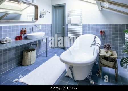 A stylish attic bathroom features blue tiles, a vintage clawfoot tub, a modern basin, and chic decor accents under a skylight. Stock Photo