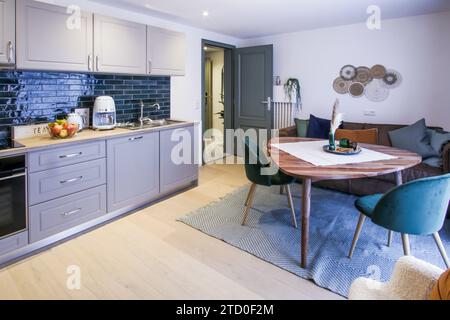 A sleek, contemporary kitchen with blue backsplash and gray cabinets, adjacent to a cozy dining space with a wooden table and blue chairs. Stock Photo