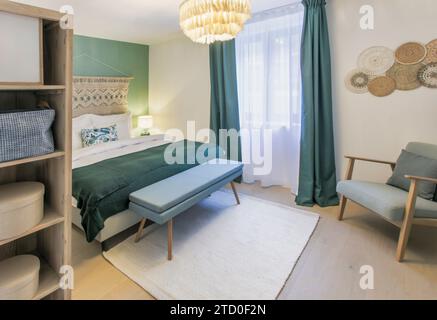 A well-lit bedroom featuring a comfortable bed with stylish pillows, a bench at the foot, a modern armchair, and decorative wall art. Stock Photo