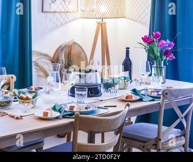An elegant dinner table is set with a modern fondue pot, wine glasses, fresh flowers, and a variety of dishes, creating a cozy dining atmosphere. Stock Photo