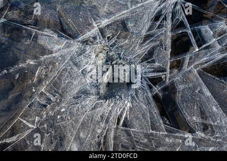 A close-up view of a frozen surface showcasing a complex network of ice cracks with a stone frozen in the center. Stock Photo