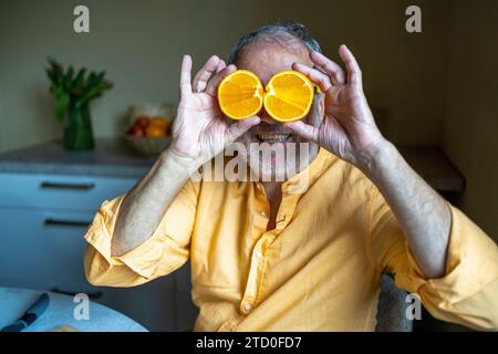 Cheerful senior man in casual clothes smiling and covering eyes with halved orange slices while sitting n kitchen Stock Photo