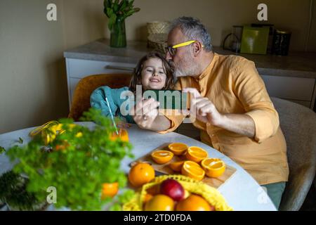 High angle view of senior grandfather kissing on happy granddaughter's forehead while taking selfie over smart phone at dining table at home Stock Photo