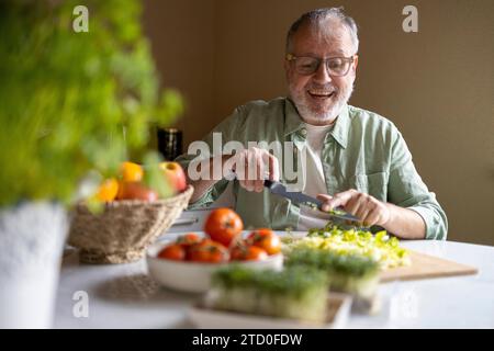 Cheerful senior man in glasses eyewear cutting lettuce in pieces with knife on wooden chopping board while preparing salad at home Stock Photo