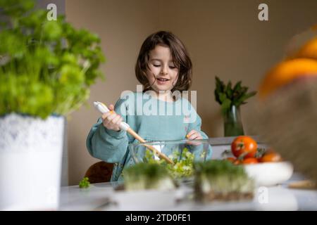 Elementary cute girl wearing casuals mixing vegetables with spatula in glass bowl while preparing salad at home Stock Photo