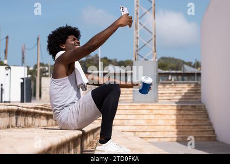 Smiling young male athlete with afro hair holding water bottle and taking selfie with smartphone while sitting on steps against blue sky after workout Stock Photo
