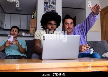 Diverse young man messaging over smartphone and friends with laptop and console playing online video game and looking away in living room during weeke Stock Photo