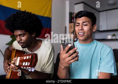 Young man with afro hair playing guitar while male friend looking at smartphone and singing song while spending weekend together at home Stock Photo