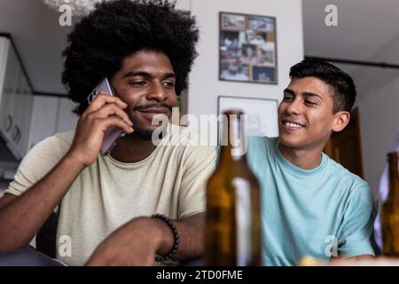 Low angle view of smiling young man looking at black friend talking over smartphone while having beer together in living room during weekend Stock Photo