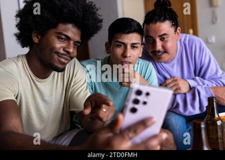 Smiling young man with afro hair taking selfie over smartphone with male friends while spending leisure time together in living room Stock Photo