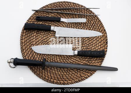 Top view of various types of knives comprising tongs bead cut chef utility and sharpening rod while placed on wicker placemat on kitchen table in dayl Stock Photo